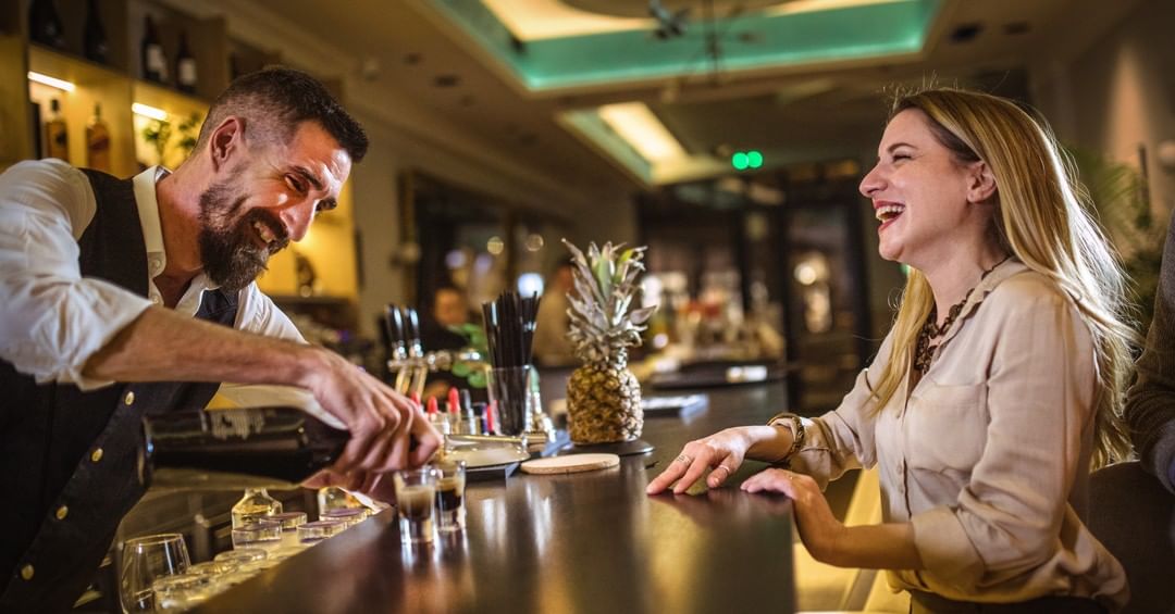 A bartender and customer laughing at the bar, he is pouring shots of drink.
